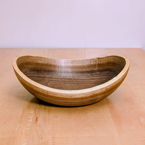 Pode incluir: Uma tigela de madeira torneada à mão com um interior castanho escuro profundo com padrões de grão de madeira visíveis e uma borda de tom natural mais claro. A tigela tem uma forma orgânica assimétrica com um acabamento liso e polido.