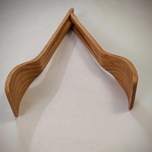 May include: Two wooden salad tongs, made from light brown wood, are interlocked at the top, forming a V-shape. The tongs have a curved design with a smooth finish, suitable for serving salads or other dishes.