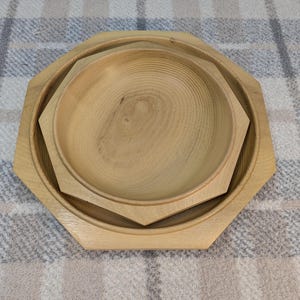 May include: A set of two wooden bowls, one nested inside the other. The bowls are light-colored, possibly unfinished wood, with a natural grain pattern visible. The outer bowl has an octagonal shape, while the inner bowl is round.