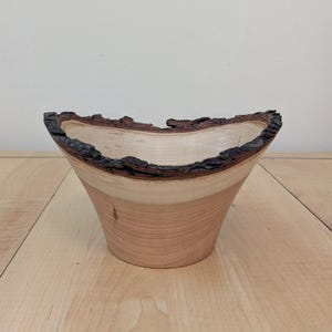 Puede incluir: Un cuenco de madera hecho a mano con un borde natural. El cuenco presenta un interior marrón claro, una banda marrón más oscura y un borde de corteza natural. El cuenco tiene forma cónica y se apoya sobre una superficie de madera.