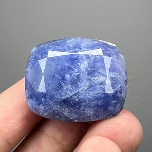May include: A faceted, square-cut gemstone with a deep blue hue. The stone has a textured appearance and a polished surface, reflecting light across its many facets. The gemstone is held in a hand.