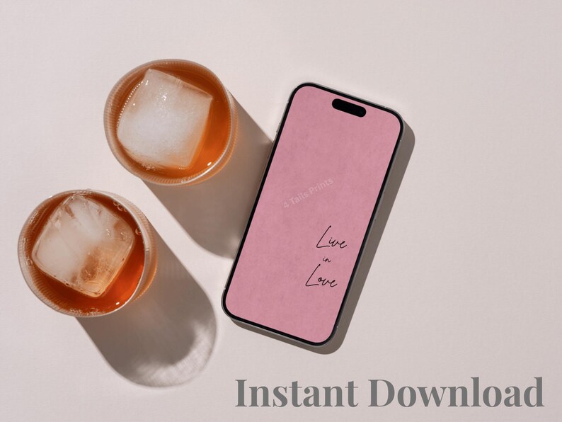 A smartphone with a pink screen displaying the text "Live in Love" and a faint "4 Tails Prints" watermark, positioned next to two round glass tumblers filled with amber-colored liquid and large square ice cubes.