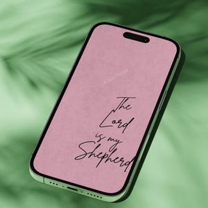 "A green iPhone mockup displaying a soft pink phone wallpaper with the handwritten-style scripture 'The Lord is My Shepherd,' photographed against a lush green tropical leaf background."