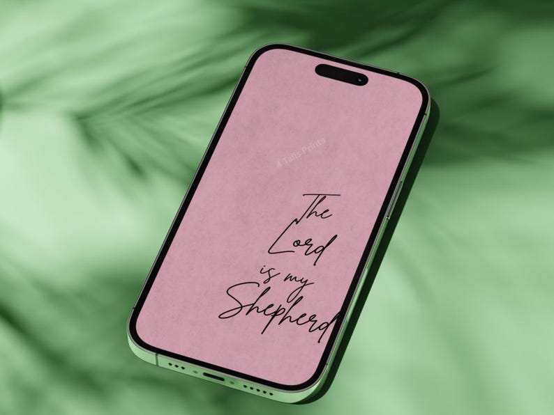 "A green iPhone mockup displaying a soft pink phone wallpaper with the handwritten-style scripture 'The Lord is My Shepherd,' photographed against a lush green tropical leaf background."
