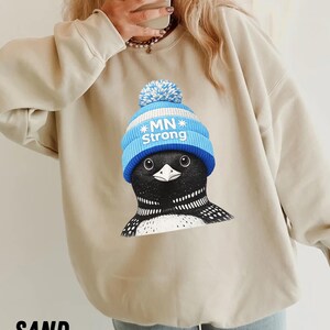 May include: Sand-coloured sweatshirt with a graphic of a loon wearing a blue knit cap with the text "MN Strong." The loon has black and white markings. The word "SAND" is printed at the bottom of the image.