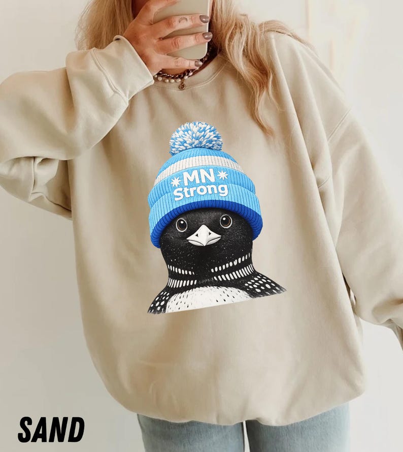 May include: Sand-coloured sweatshirt with a graphic of a loon wearing a blue knit cap with the text "MN Strong." The loon has black and white markings. The word "SAND" is printed at the bottom of the image.