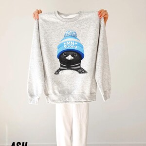 May include: Ash grey sweatshirt with a cartoon loon wearing a blue knit cap with the text "MN Strong". The loon has black and white markings. The sweatshirt is held up against a white background.