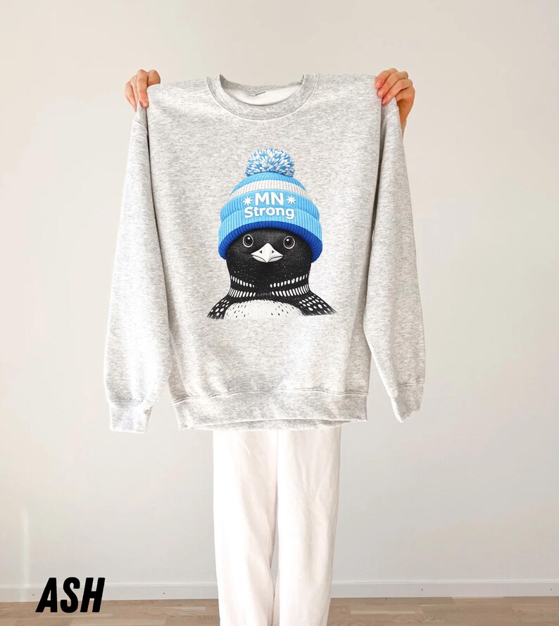May include: Ash grey sweatshirt with a cartoon loon wearing a blue knit cap with the text "MN Strong". The loon has black and white markings. The sweatshirt is held up against a white background.