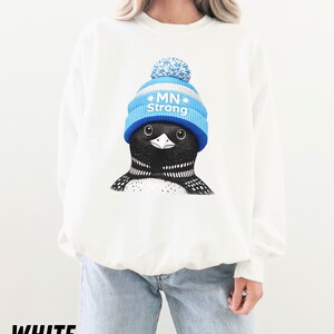 May include: White sweatshirt with a graphic of a loon wearing a blue and white knitted hat with the text "MN Strong." The loon has a black and white patterned chest and a black beak. The word "WHITE" is printed at the bottom.