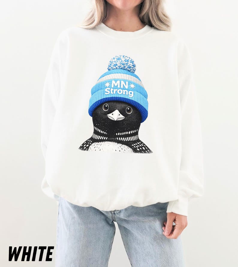 May include: White sweatshirt with a graphic of a loon wearing a blue and white knitted hat with the text "MN Strong." The loon has a black and white patterned chest and a black beak. The word "WHITE" is printed at the bottom.