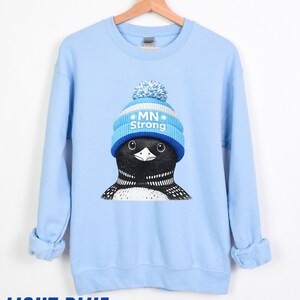 May include: Pale blue sweatshirt with a graphic of a loon wearing a blue and white knitted hat with the text "MN Strong." The loon has black and white feathers. The words "LIGHT BLUE" are printed at the bottom. The sweatshirt is hanging on a wooden hanger.