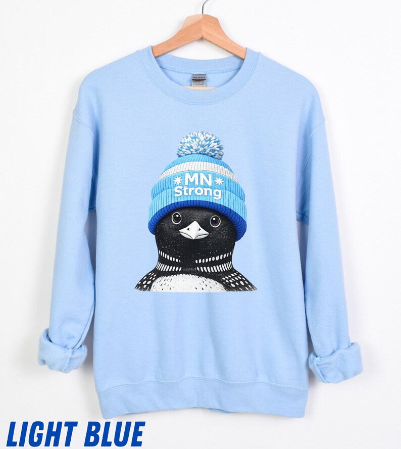 May include: Pale blue sweatshirt with a graphic of a loon wearing a blue and white knitted hat with the text "MN Strong." The loon has black and white feathers. The words "LIGHT BLUE" are printed at the bottom. The sweatshirt is hanging on a wooden hanger.
