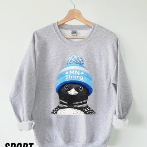 May include: A grey sweatshirt with a graphic of a loon wearing a blue and white knitted hat with the text "MN Strong." The sweatshirt has long sleeves and a crew neck. The word "SPORT" is printed in black at the bottom.