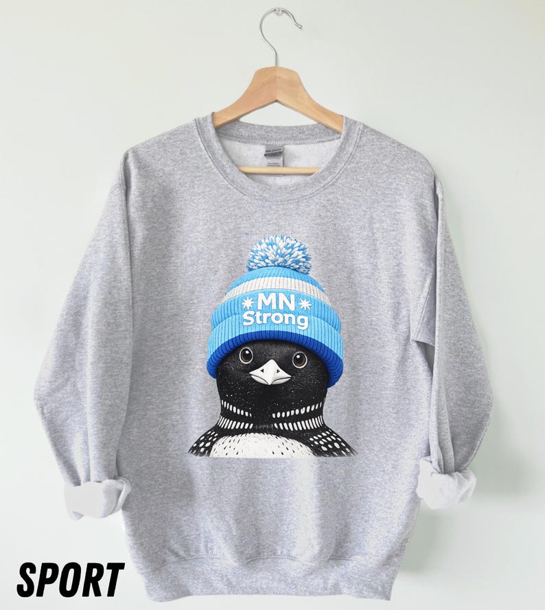May include: A grey sweatshirt with a graphic of a loon wearing a blue and white knitted hat with the text "MN Strong." The sweatshirt has long sleeves and a crew neck. The word "SPORT" is printed in black at the bottom.