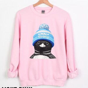 May include: Pale pink sweatshirt with a graphic of a loon wearing a blue and white knitted hat with the text "MN Strong". The sweatshirt has long sleeves and a crew neck. The text "LIGHT PINK" is at the bottom.