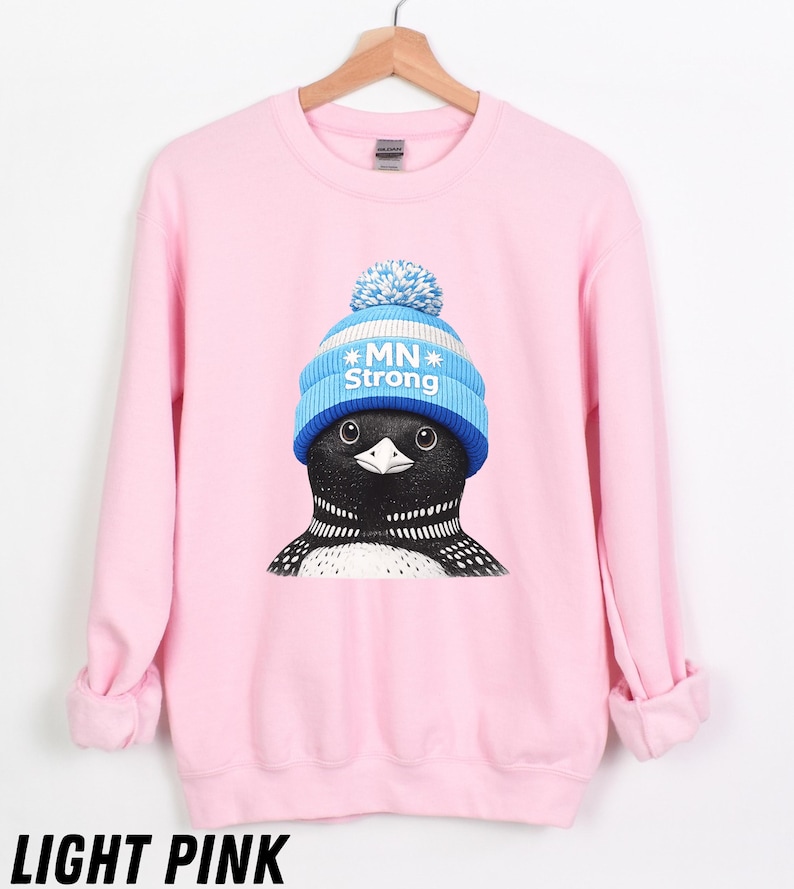 May include: Pale pink sweatshirt with a graphic of a loon wearing a blue and white knitted hat with the text "MN Strong". The sweatshirt has long sleeves and a crew neck. The text "LIGHT PINK" is at the bottom.