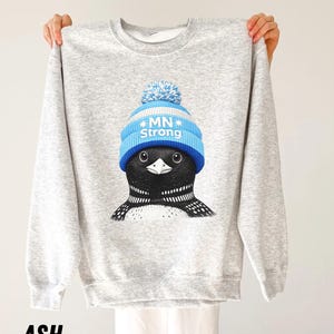 May include: A light grey sweatshirt with a graphic of a loon wearing a blue and white knitted hat with the text "MN Strong". The sweatshirt is labelled "ASH" at the bottom.
