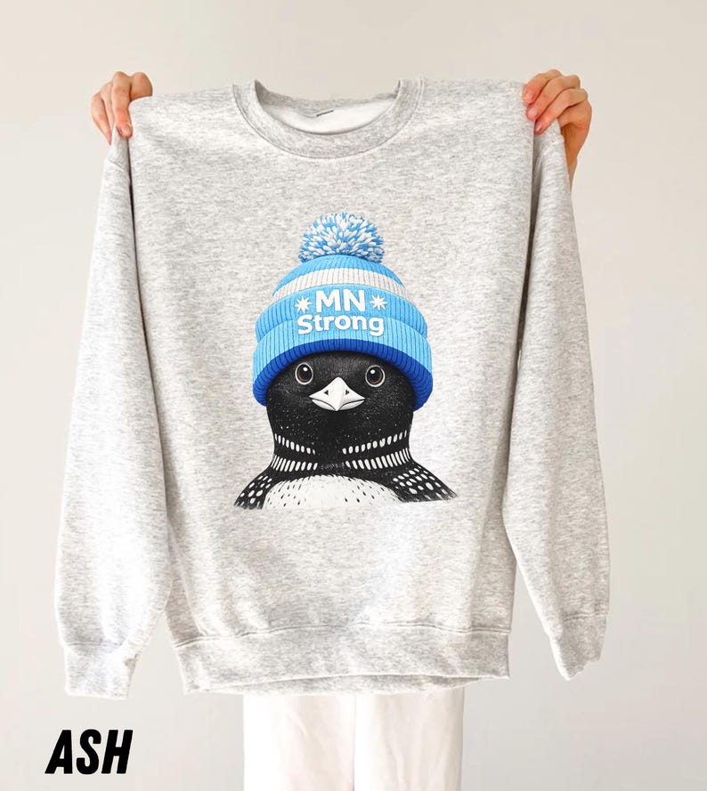 May include: A light grey sweatshirt with a graphic of a loon wearing a blue and white knitted hat with the text "MN Strong". The sweatshirt is labelled "ASH" at the bottom.