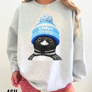 May include: A light grey sweatshirt with a graphic of a black and white bird wearing a blue and white knitted hat with the text "MN Strong". The word "ASH" is printed at the bottom.