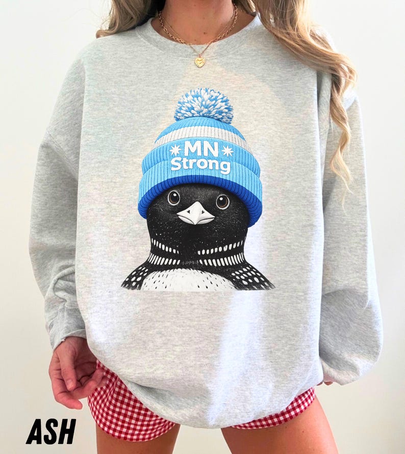 May include: A light grey sweatshirt with a graphic of a black and white bird wearing a blue and white knitted hat with the text "MN Strong". The word "ASH" is printed at the bottom.