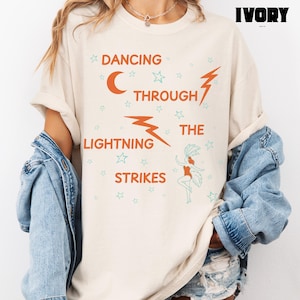 Pode incluir: Camiseta cor marfim com o texto "DANCING THROUGH THE LIGHTNING STRIKES" em laranja e verde-azulado. O design inclui uma lua crescente, raios, estrelas e uma ilustração de bailarina. Roupa casual.