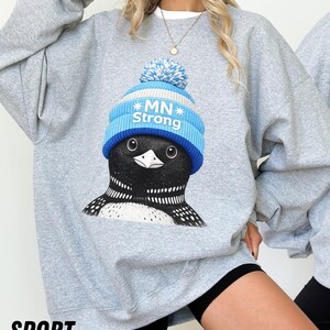 May include: A light grey sweatshirt with a graphic of a loon wearing a blue and white knit hat with the text "MN Strong." The sweatshirt has a crew neck and long sleeves. The word "SPORT" is printed in black at the bottom.