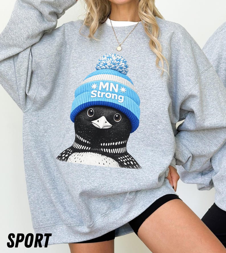 May include: A light grey sweatshirt with a graphic of a loon wearing a blue and white knit hat with the text "MN Strong." The sweatshirt has a crew neck and long sleeves. The word "SPORT" is printed in black at the bottom.