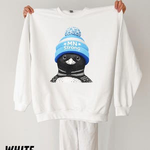 May include: White sweatshirt with a graphic of a loon wearing a blue and white knit hat with the text "MN Strong." The loon has black and white feathers and is centered on the front. The word "WHITE" is printed at the bottom.