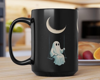 May include: Black ceramic mug featuring a whimsical illustration of a ghost on a swing under a crescent moon. The ghost is draped in a white sheet, and the background is a starry night sky. The mug has a large handle.