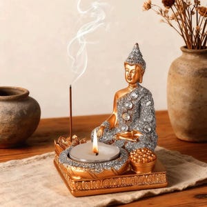 May include: A gold and silver Buddha statue with a lit candle and incense. The statue is seated in a meditative pose, with intricate detailing on the robe and base. A small bowl of gold beads sits next to the candle. The incense stick is emitting smoke.