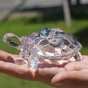 May include: A clear, crystal turtle figurine with a detailed shell and legs. The turtle is transparent, allowing light to pass through, and is held in a person's hand against a blurred green background. The figurine is a decorative item.