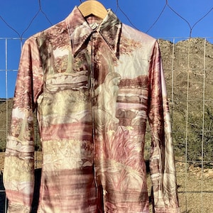 May include: A long-sleeve button-down shirt featuring a continuous, muted pink and cream-toned landscape print. The design depicts a seated figure amidst trees and foliage, with a pointed collar, buttoned cuffs, and a central vertical button placket.
