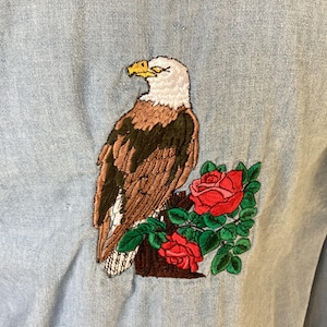 May include: Light blue denim shirt with an embroidered design of a bald eagle perched on a branch with red roses and green leaves. The eagle has brown and white feathers and a yellow beak.