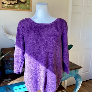 May include: A purple long-sleeved sweater with a textured, speckled knit pattern. The garment features a wide, ribbed neckline and a straight hemline, with small buttons visible along the shoulder seams on both sides of the collar.