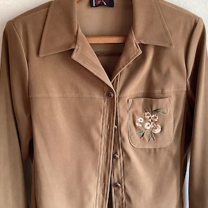 May include: A tan long-sleeve button-down shirt featuring a pointed collar and a single chest pocket. The pocket is decorated with an embroidered floral design in shades of pink, white, and green. The shirt has a front button closure.