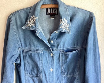 May include: A light blue denim shirt featuring a pointed collar with white floral embroidery on both sides. The shirt has a button-down front, long sleeves, and two chest pockets. A black label at the inner neckline displays the text AT LAST.