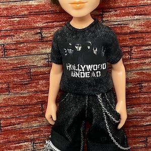 Könnte beinhalten: Eine Puppe mit schwarzer Mütze, einem schwarzen T-Shirt mit dem Aufdruck "HOLLYWOOD UNDEAD" und einer schwarzen Jeanshose mit silbernen Reißverschlüssen und einer Kette. Die Puppe trägt braune Schuhe.