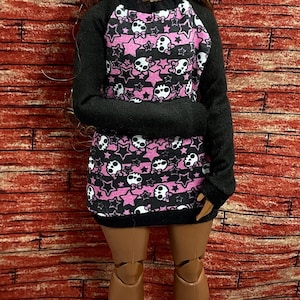 May include: A doll wearing a black and pink patterned dress with a skull and star design. The doll has brown hair and is wearing black high-heeled boots with gold accents. The background is a red brick wall.