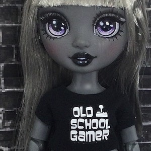 K&ouml;nnte beinhalten: Eine schwarz-wei&szlig;e Puppe mit langen grauen Haaren und einem schwarzen Shirt, auf dem in wei&szlig;en Buchstaben "Old School Gamer" steht. Die Puppe hat violette Augen und ein schwarz-wei&szlig;es Outfit.