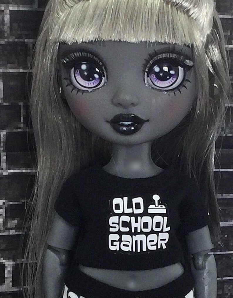 K&ouml;nnte beinhalten: Eine schwarz-wei&szlig;e Puppe mit langen grauen Haaren und einem schwarzen Shirt, auf dem in wei&szlig;en Buchstaben "Old School Gamer" steht. Die Puppe hat violette Augen und ein schwarz-wei&szlig;es Outfit.