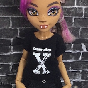 K&ouml;nnte beinhalten: Ein schwarzes T-Shirt mit einem wei&szlig;en "Generation X" Aufdruck auf der Vorderseite. Die Puppe tr&auml;gt das Shirt und hat pink- und lilafarbenes Haar.