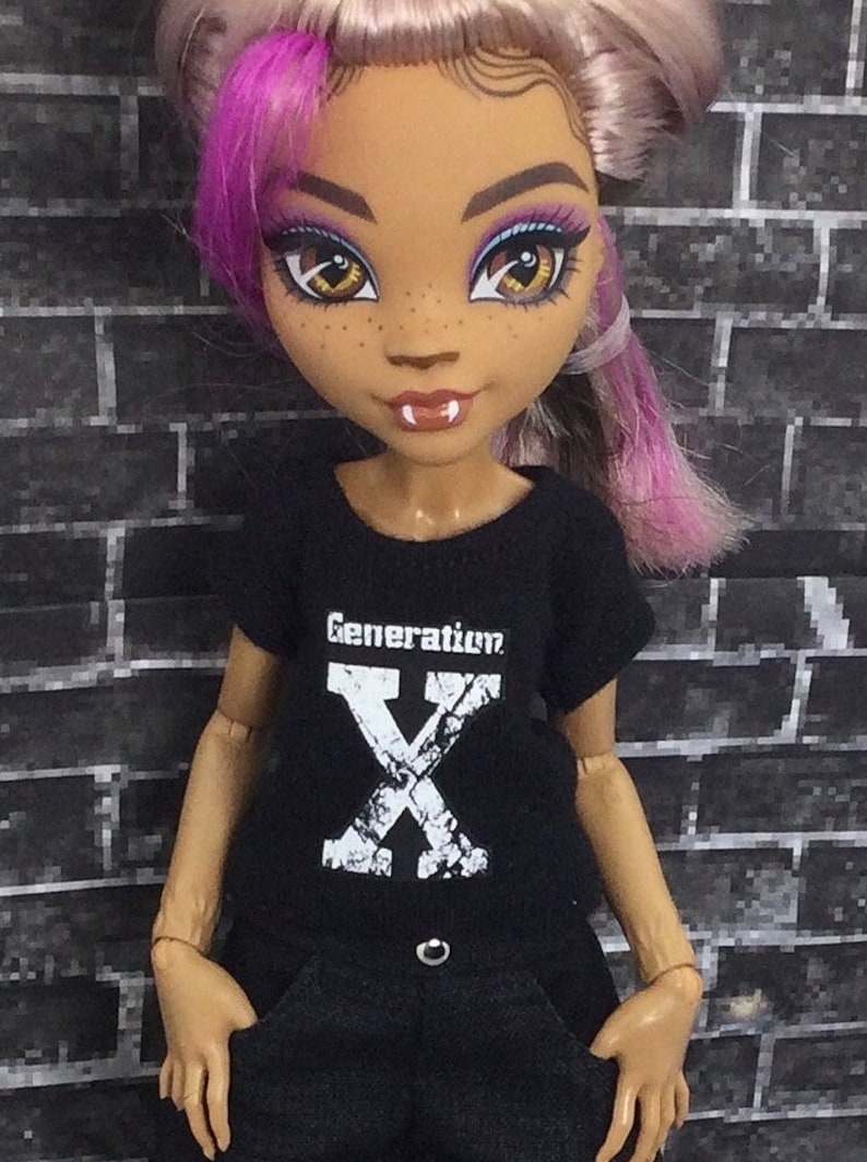 K&ouml;nnte beinhalten: Ein schwarzes T-Shirt mit einem wei&szlig;en "Generation X" Aufdruck auf der Vorderseite. Die Puppe tr&auml;gt das Shirt und hat pink- und lilafarbenes Haar.