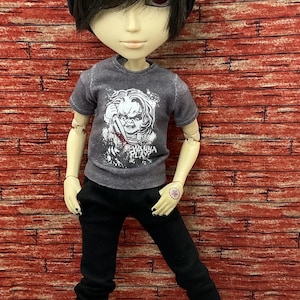May include: A doll wearing a black beanie, a gray t-shirt with a graphic of a character holding a knife, black pants, and white sneakers with pink soles. The text "Wanna Play?" is on the shirt.