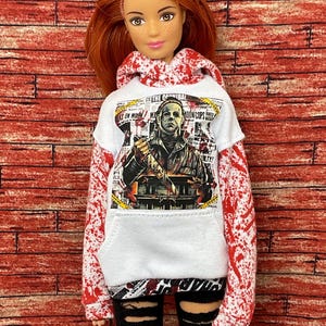 May include: A doll with red hair wearing a white hoodie with a graphic of Michael Myers from the movie "Halloween". The hoodie has red splatter paint on the sleeves and hood. The doll is also wearing black ripped leggings.