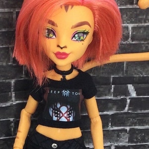 K&ouml;nnte beinhalten: Eine Puppe mit orangefarbenem und rosafarbenem Haar, die ein schwarzes Crop-Top mit einem Band-Logo tr&auml;gt, auf dem "SLEEP TOKE" steht, und schwarze Jeans-Shorts.