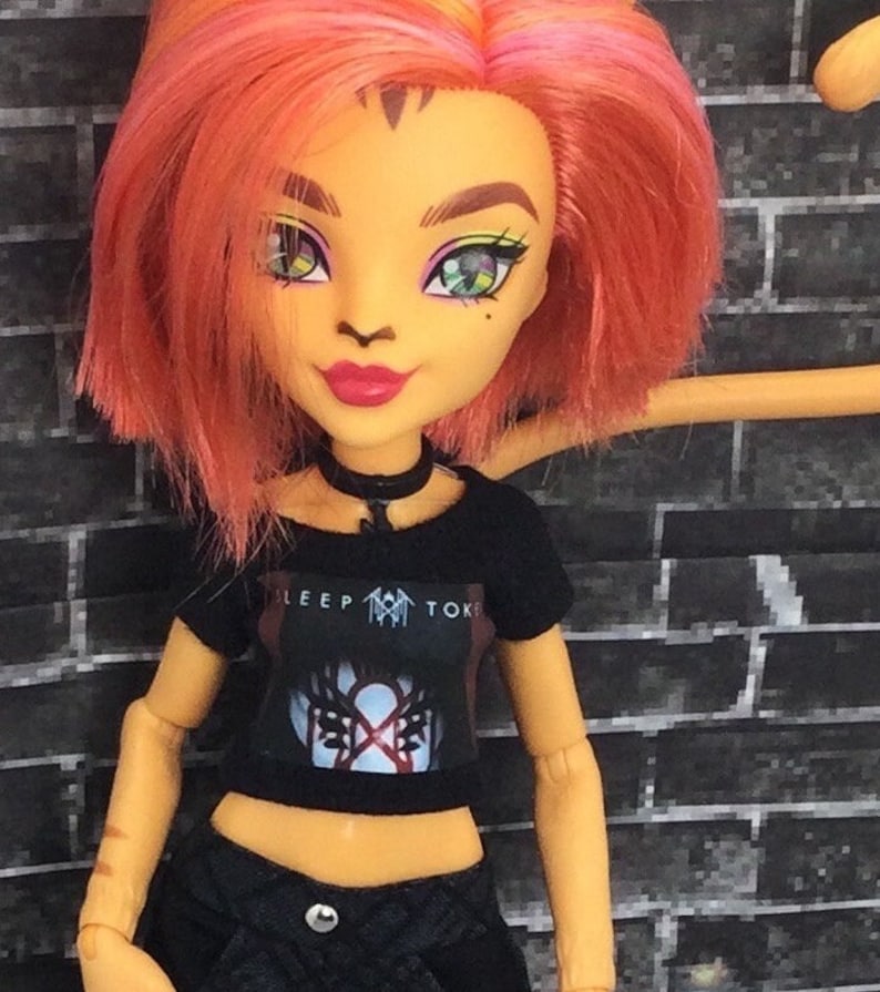 K&ouml;nnte beinhalten: Eine Puppe mit orangefarbenem und rosafarbenem Haar, die ein schwarzes Crop-Top mit einem Band-Logo tr&auml;gt, auf dem "SLEEP TOKE" steht, und schwarze Jeans-Shorts.