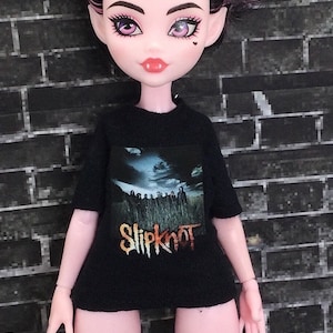K&ouml;nnte beinhalten: Ein schwarzes T-Shirt mit einem grafischen Aufdruck der Band Slipknot. Die Bandmitglieder sind in einem Feld mit einem bew&ouml;lkten Himmel im Hintergrund abgebildet. Der Name der Band ist in orangefarbenen und gelben Buchstaben unter dem Bild gedruckt.