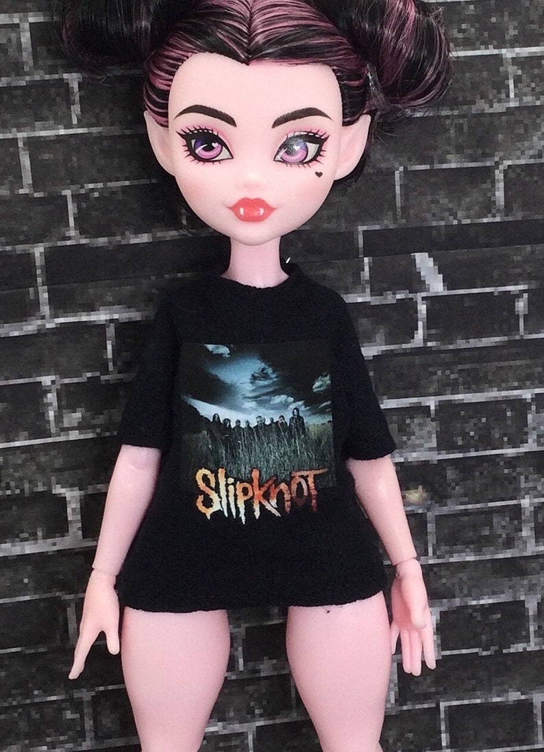 K&ouml;nnte beinhalten: Ein schwarzes T-Shirt mit einem grafischen Aufdruck der Band Slipknot. Die Bandmitglieder sind in einem Feld mit einem bew&ouml;lkten Himmel im Hintergrund abgebildet. Der Name der Band ist in orangefarbenen und gelben Buchstaben unter dem Bild gedruckt.