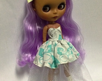 May include: A doll with long, wavy purple hair, wearing a white dress with a butterfly print and a white tulle skirt. The doll has brown skin and is wearing white boots.