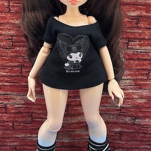Könnte beinhalten: Eine Puppe mit langem braunen Haar in zwei Zöpfen, bekleidet mit einem schwarzen schulterfreien T-Shirt mit einer Grafik der Figur Kuromi und dem darunter gedruckten Text KUROMI. Die Puppe trägt schwarz-weiß gestreifte Stulpen und schwarze Schuhe.